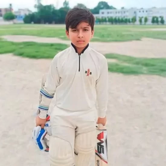 Young Vaibhav Sooryavanshi stands in white cricket kit holding a bat and gloves on a local ground.